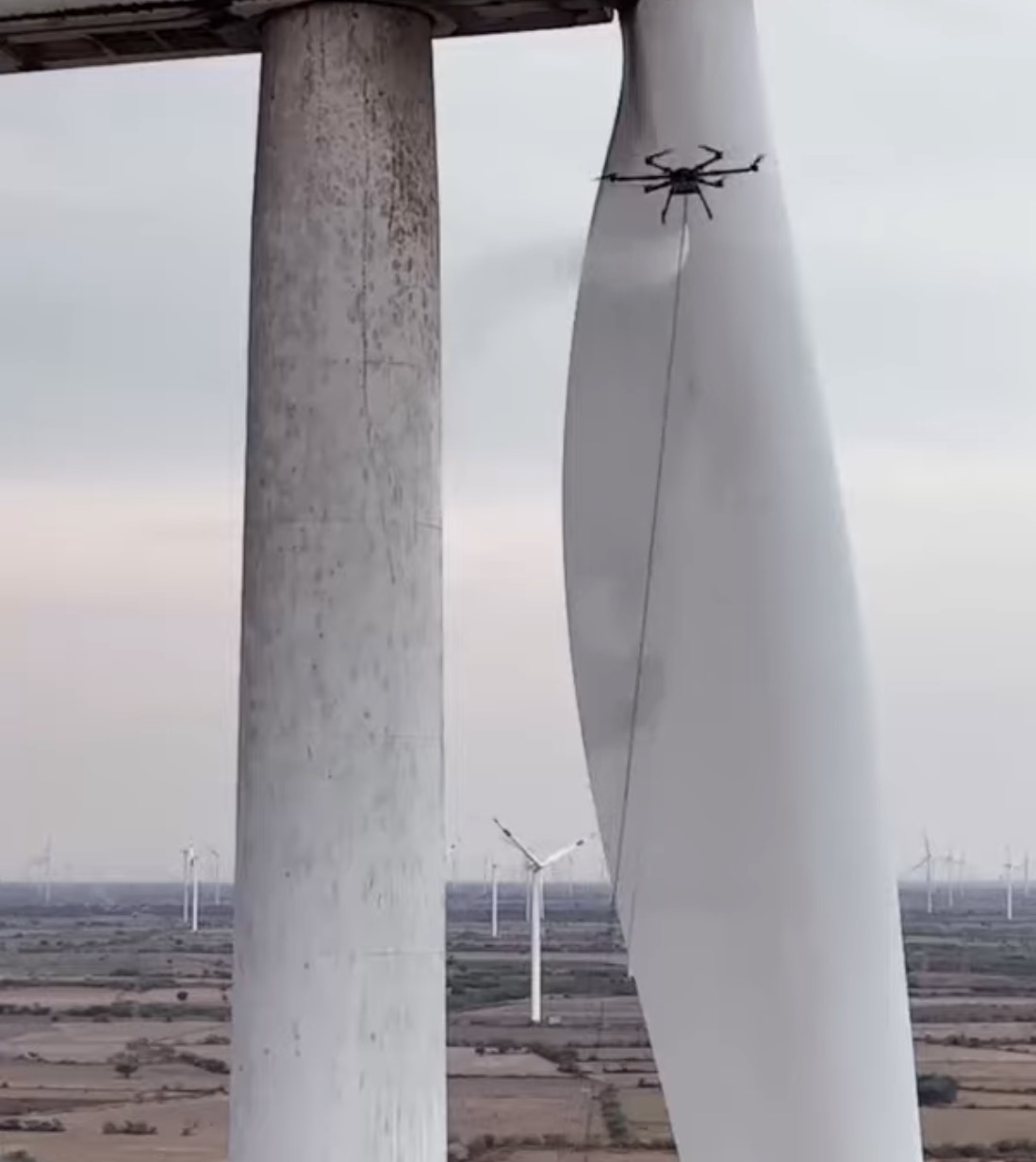 StratoClean drone cleaning a wind-turbine blade high on the tower with a wind farm behind — touchless, no rope-access crews, no turbine downtime for scaffolding